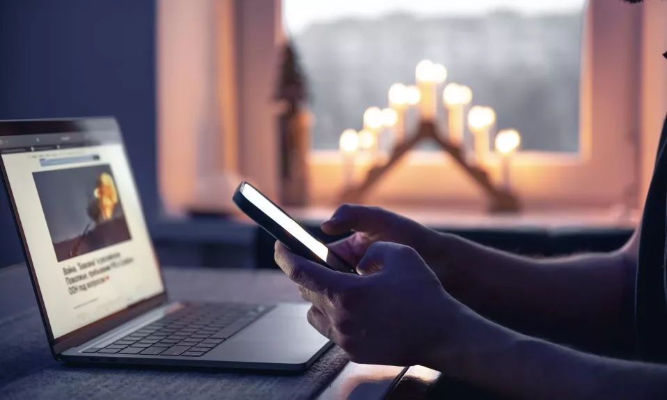 Person using smartphone by candlelit window with laptop.
